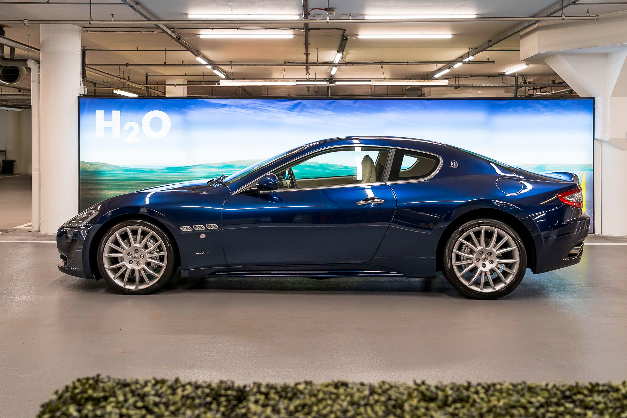 Side view of a dark blue Maserati coupe parked in front of an H2O backdrop