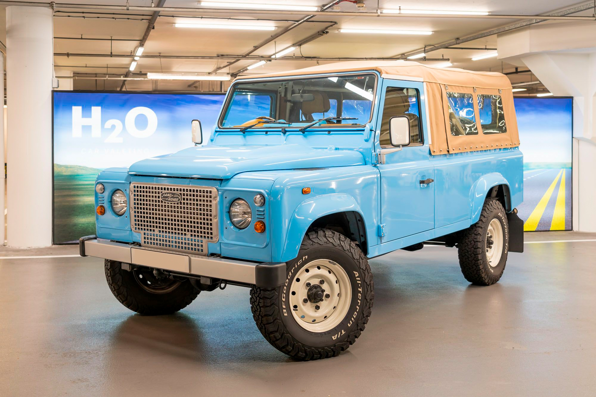 Light blue vintage Land Rover Defender with tan soft top parked in front of an H2O backdrop