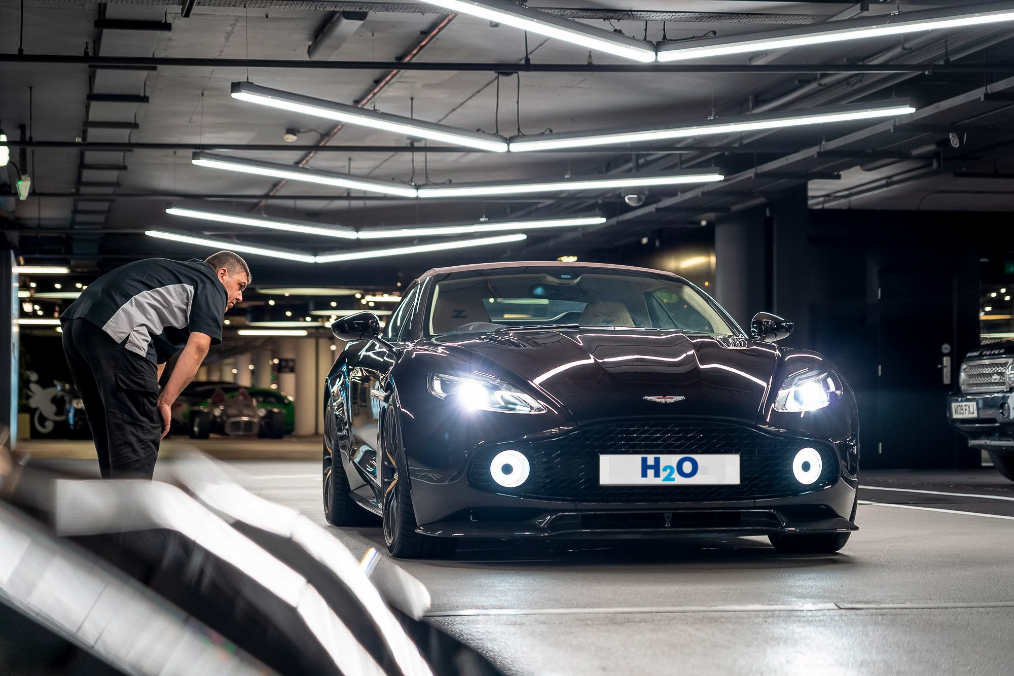 Valeter in a grey and black uniform inspecting a black Aston Martin with headlights on in a car park